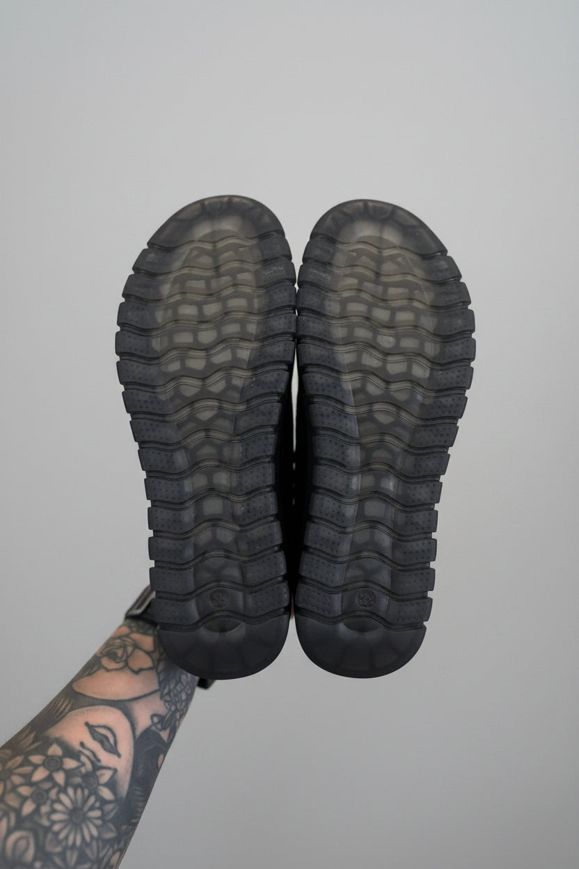 Black boots with tread pattern held by a person with tattoos on a light surface.