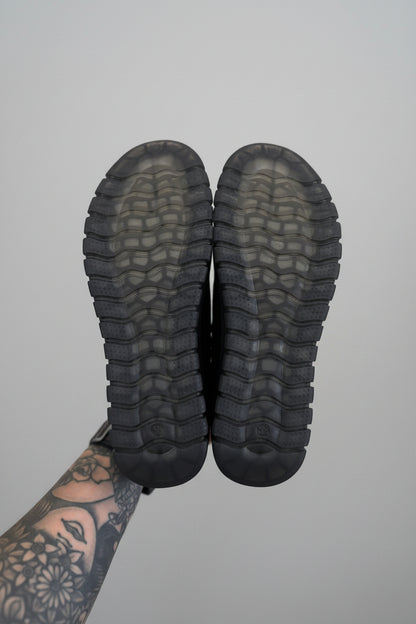 Black boots with tread pattern held by a person with tattoos on a light surface.